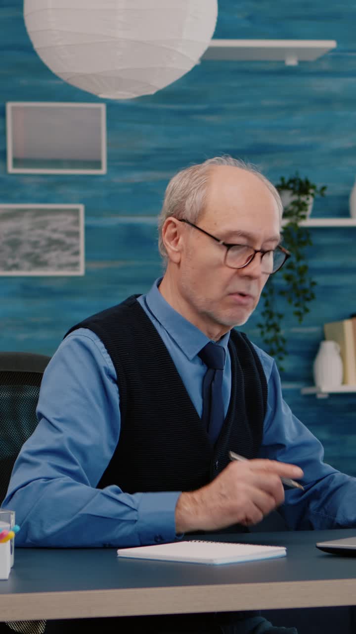 Vertical video: Multitasking retired man reading on laptop and writing on notebook