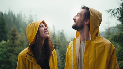 una pareja feliz: un chico y una chica con chaquetas amarillas de pie contra el telón de fondo de un bosque de montaña brumoso, sonriendo y esperando la próxima lluvia. chico y chica miran al cielo y se abrazan