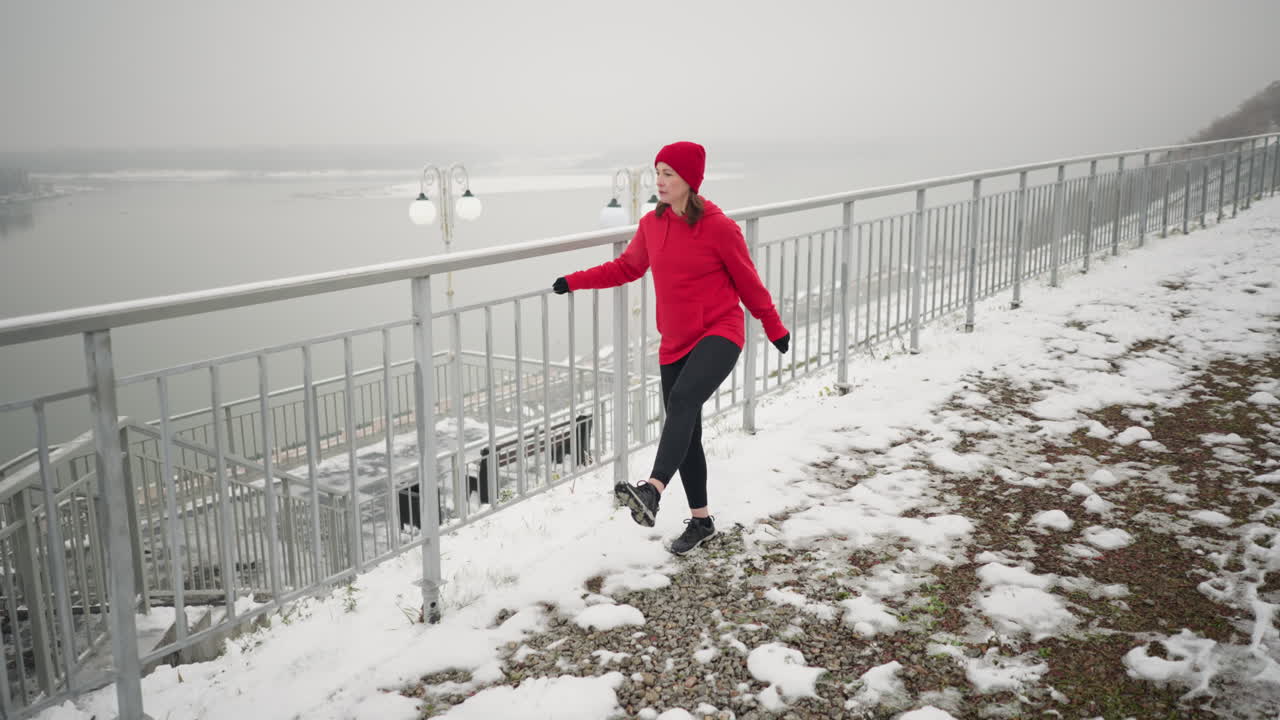 겨울 동안 야외에서 운동하는 여성, 균형을 잡기 위해 철의 레일링을 들고 다리를 앞뒤로 는 동안 안개가 많은 대기, 멀리 떨어진 강, 조명 기둥, 눈 인 땅