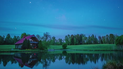 Toma-De-Tiempo-Del-Movimiento-Estelar-A-Lo-Largo-Del-Cielo-Nocturno-Sobre-Una-Pequeña-Casa-Al-Lado-De-Un-Lago