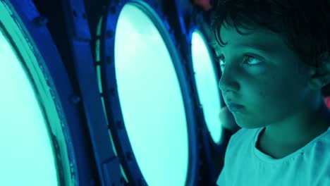 a young boy looks out the round windows of a submarine with wonder in his eyes