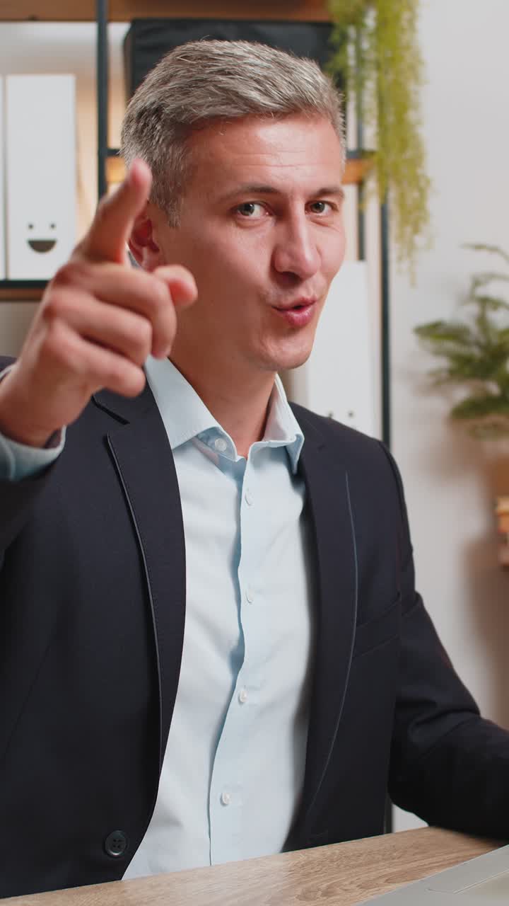 joven empresario caucásico feliz en traje con portátil apuntando a la cámara sentado en el escritorio de la oficina de casa