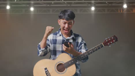 asian teenager with guitar using smartphone and celebrating the succeed while standing on stage