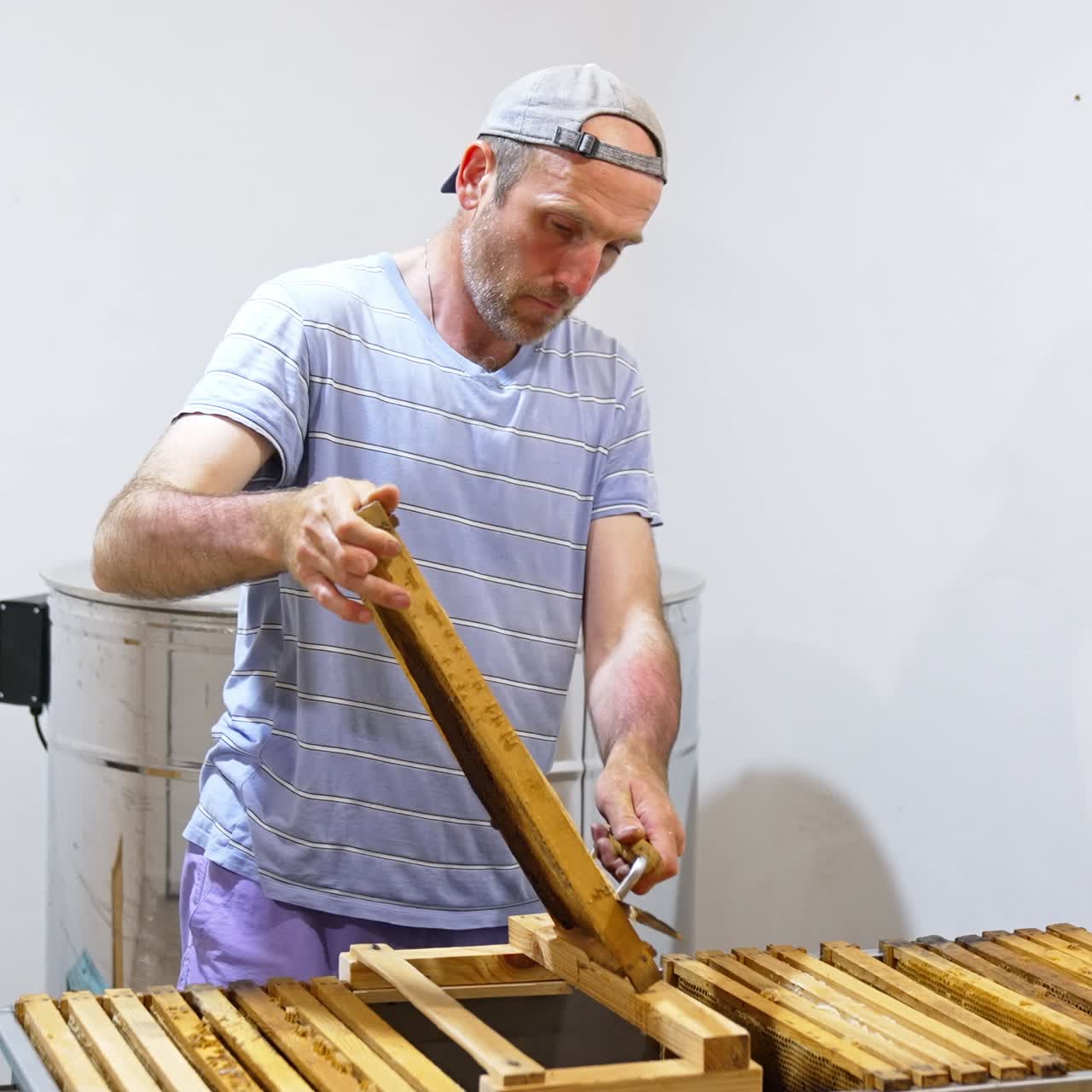 Bearded man in cap pulls a frame from a big metal box. Apiarist applies electric knife to open cells on the honey frame to prepare it for extraction