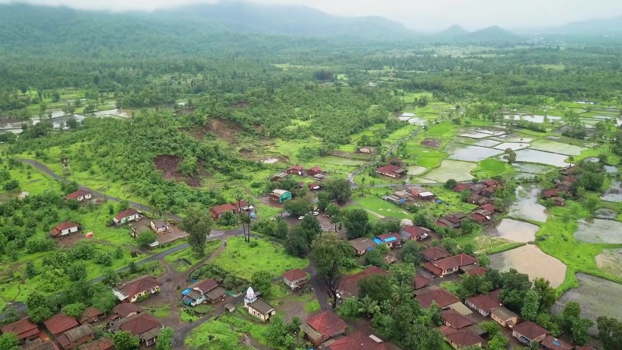 Desa pedesaan yang terletak di antara lahan pertanian hijau subur dengan sawah dan pepohonan yang tersebar selama musim hujan. Rekaman drone udara yang mulus menampilkan pemandangan pertanian dengan pegunungan berkabut.