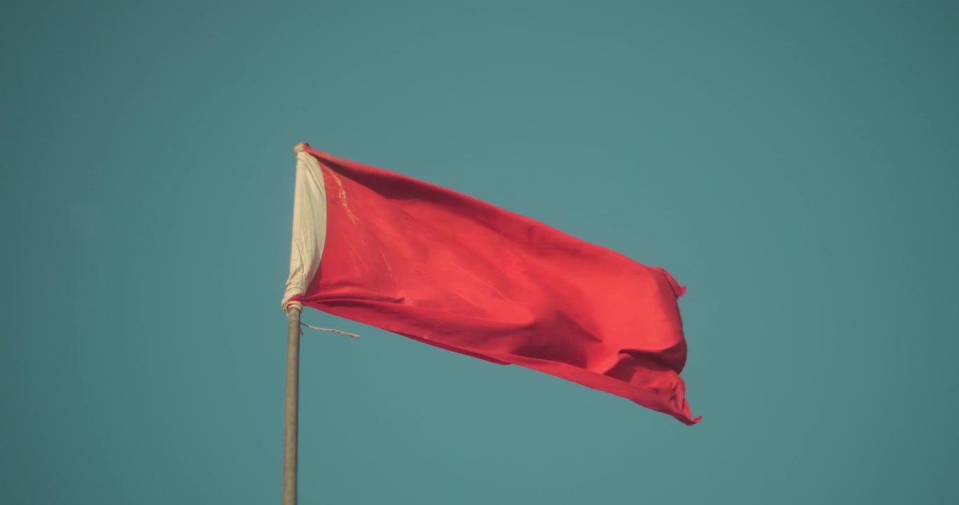 Bendera peringatan merah berkibar ditiup angin pantai yang kencang dengan latar langit biru. Simbol keselamatan pantai ini menandakan kondisi berenang yang berbahaya dan cuaca laut yang buruk.