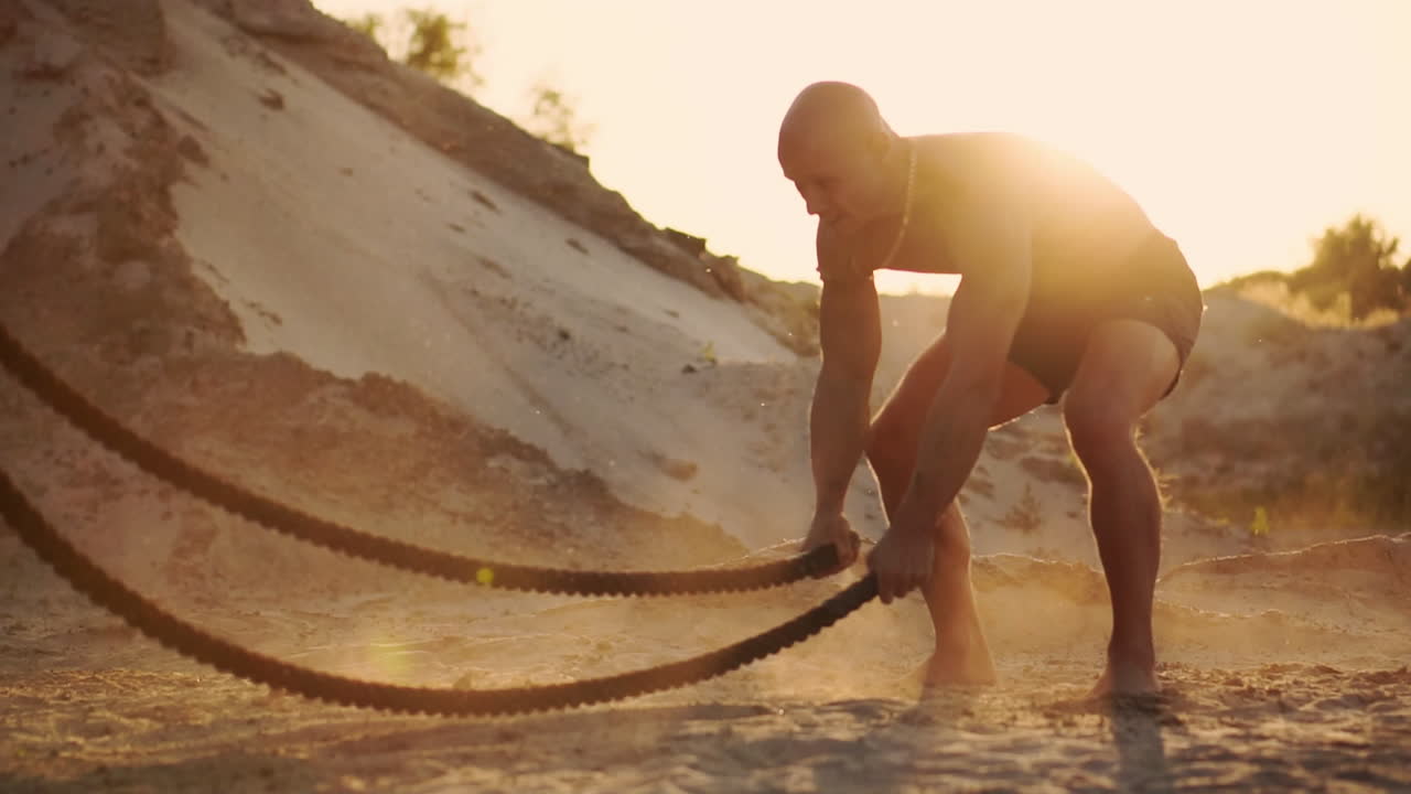atleta masculino haciendo flexiones en la playa y golpeando la cuerda en el suelo, entrenamiento circular en el sol en una playa de arena levantando polvo en cámara lenta.