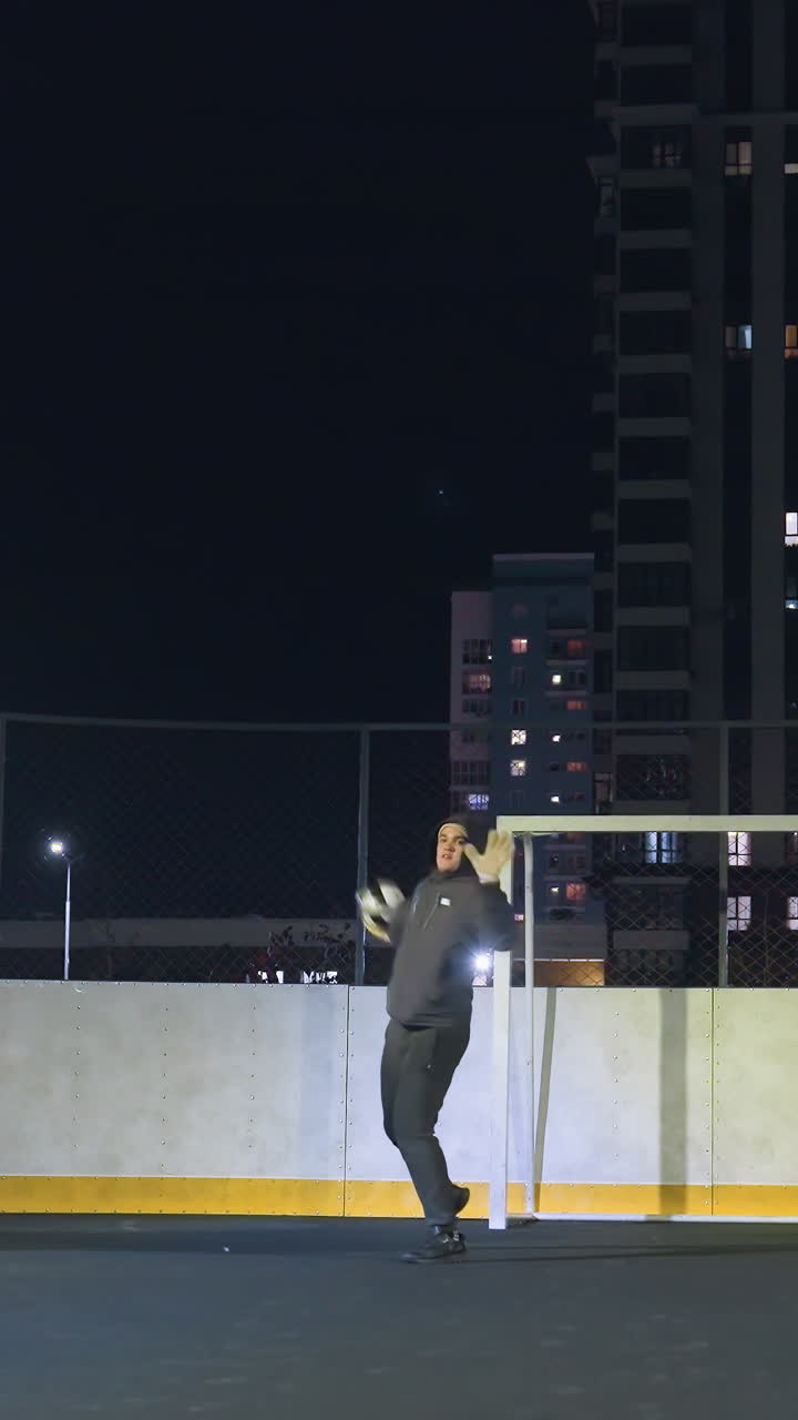 Athlete prepares to throw soccer ball during night practice near goalpost on illuminated urban field, player demonstrates athletic form under stadium lighting with residential buildings in background
