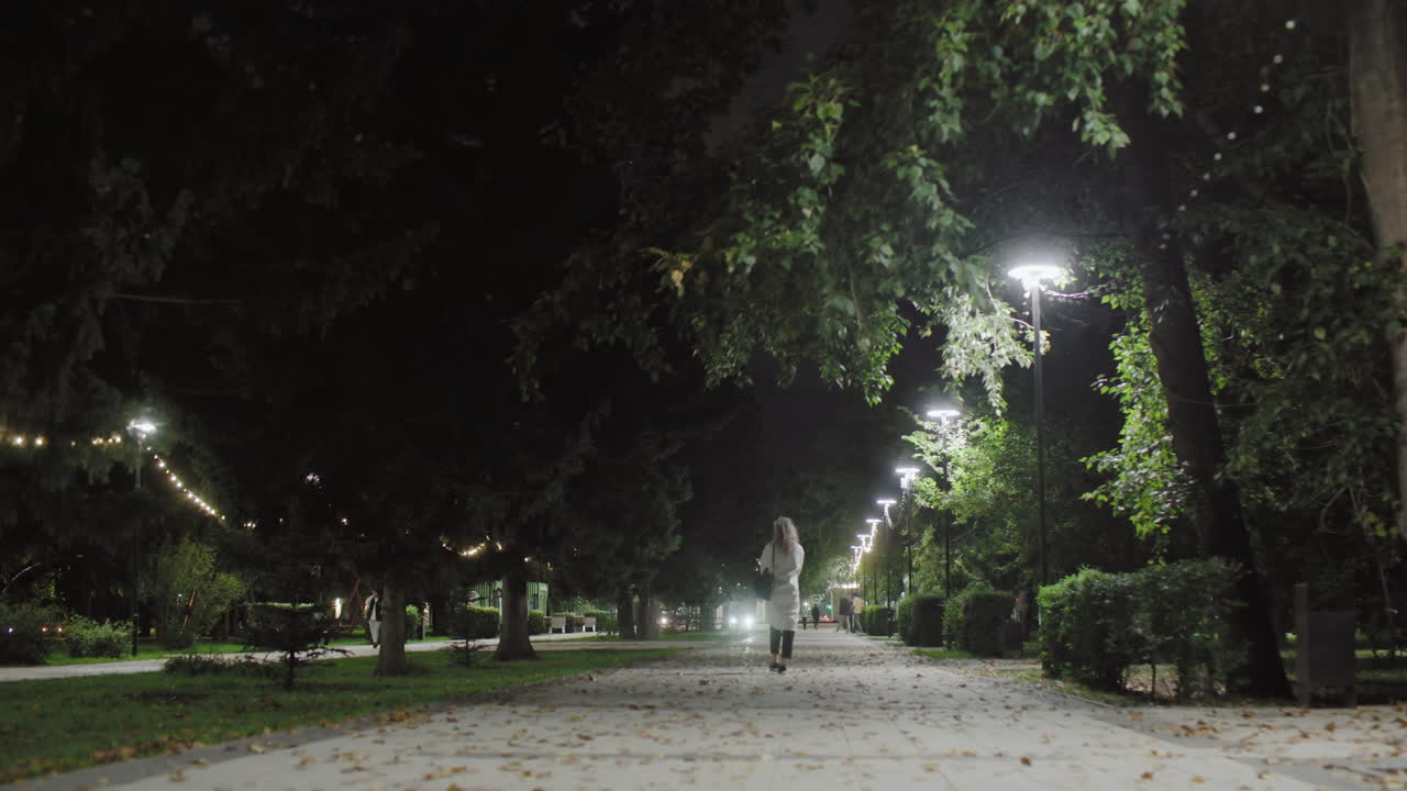 Woman in white coat walks along decorated park path at night, surrounded by trees, street lamps casting warm glow, dry leaves scattered underfoot, with people and car headlights glowing in distance