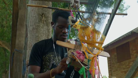 happy african artisan man weaving kente textile on rustic loom while listening music