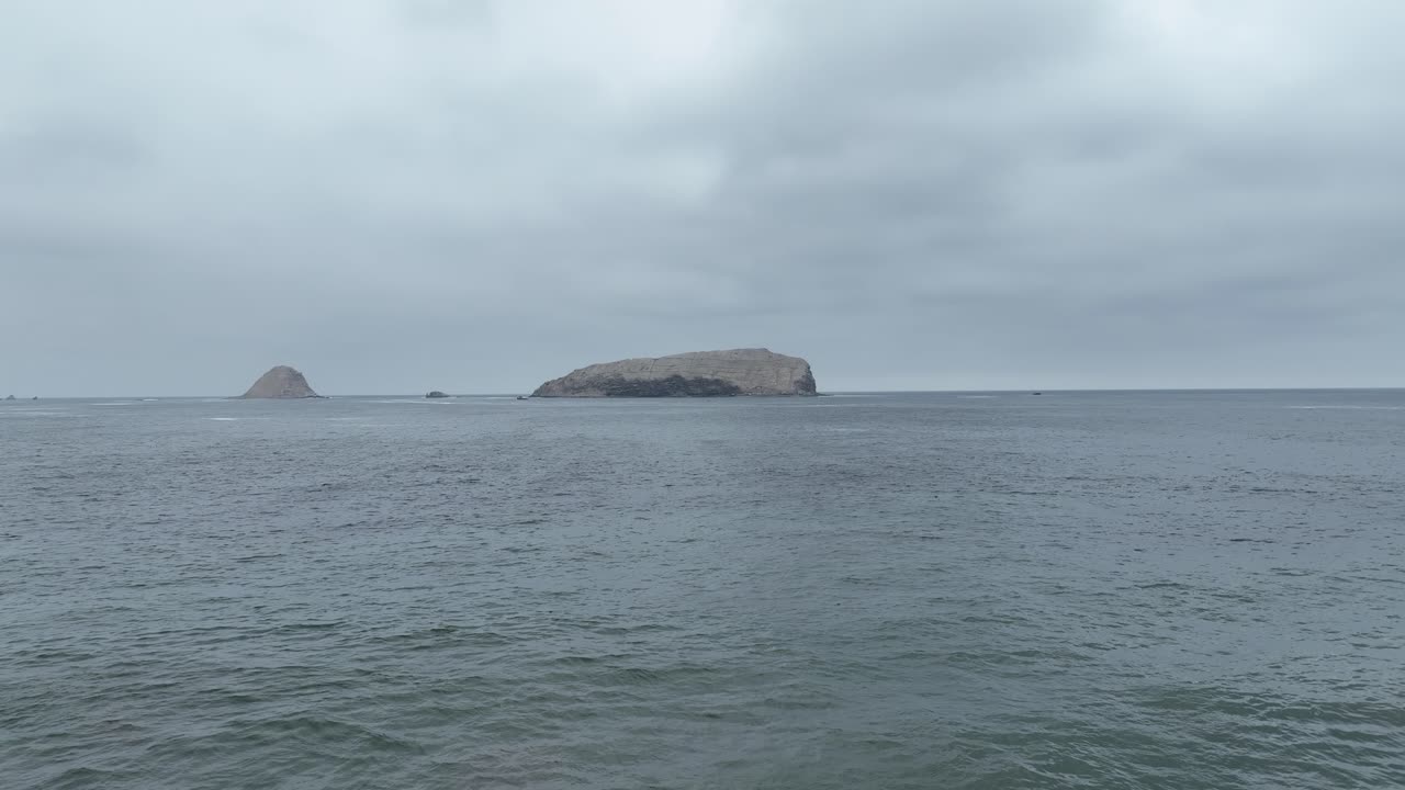페루 리마의 푸쿠수나 해변에서 바다 위의 바위와 보트가 있는 섬의 전망을 배경으로 공중 낮은 돌리 포워드 샷