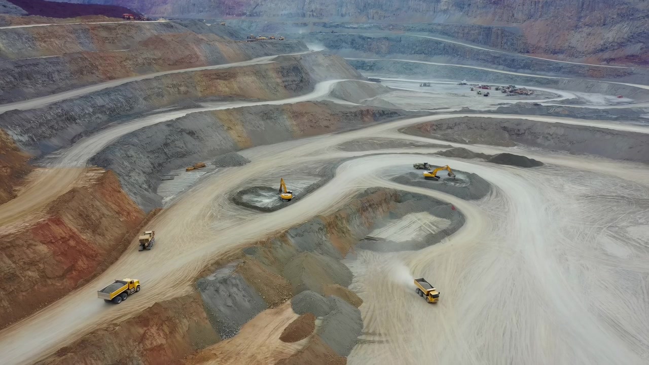 High oblique aerial view of expansive openpit mine showing hydraulic excavators loading material while articulated dump trucks move along curved haul roads beside terraced rock walls and dusty work