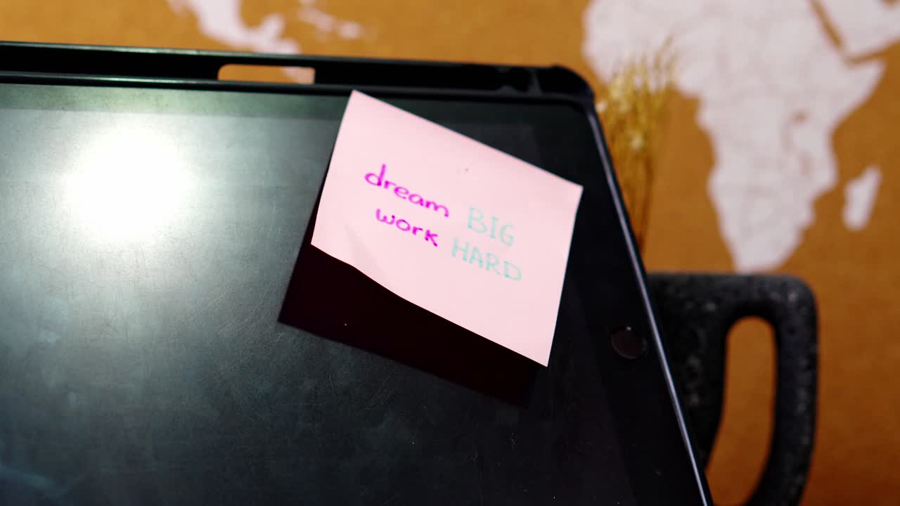 Close-up of hand removes pink sticky note saying "dream BIG work HARD" from black tablet screen