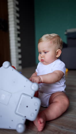 lovely serious baby boy in white romper sitting on the floor. cute child plays with toy. vertical video.