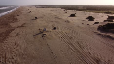 Aerial-shot-of-four-wheel-vehicle-driving-on-sandy-beach-tour-during-sunset-at-horizon---Mar-de-las-Pampas-in-Argentina