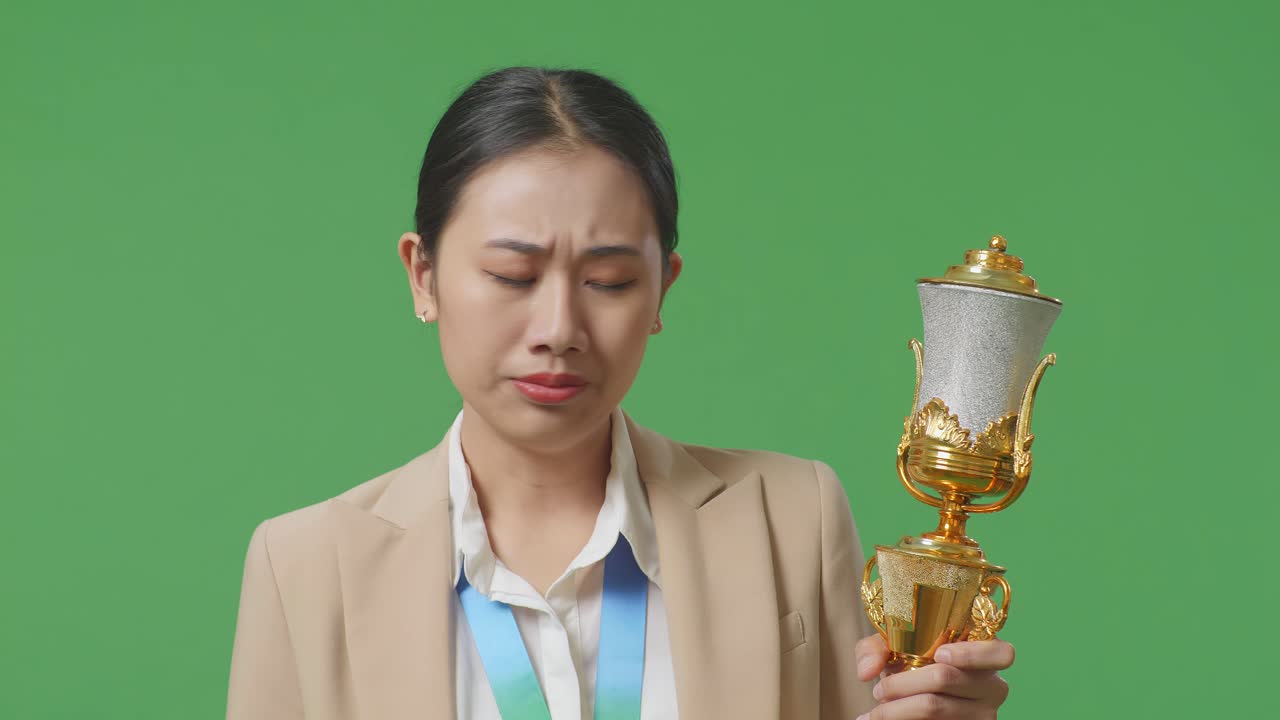 화가 난 아시안 사업가 여성은 금메달을  슈트를 입고 트로피를 보고 머리를 흔들며 불만을 표하며 스튜디오의 녹색 스크린 배경에서 첫 번째 우승자로 승리합니다.