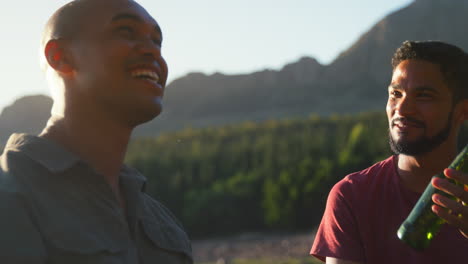 two male friends relaxing in countryside by lake and mountains drinking beer and doing cheers