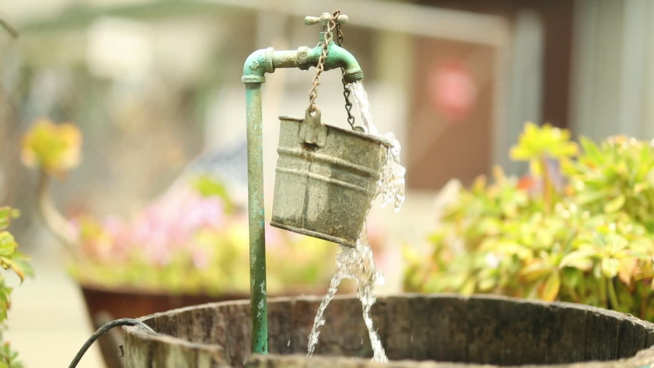 Water from a garden spout pours and spills over a hanging bucket into a larger barrel