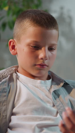 schoolboy finishes phone call looking at screen of gadget. serious boy holds smartphone in hand turning to camera while sitting on sofa closeup