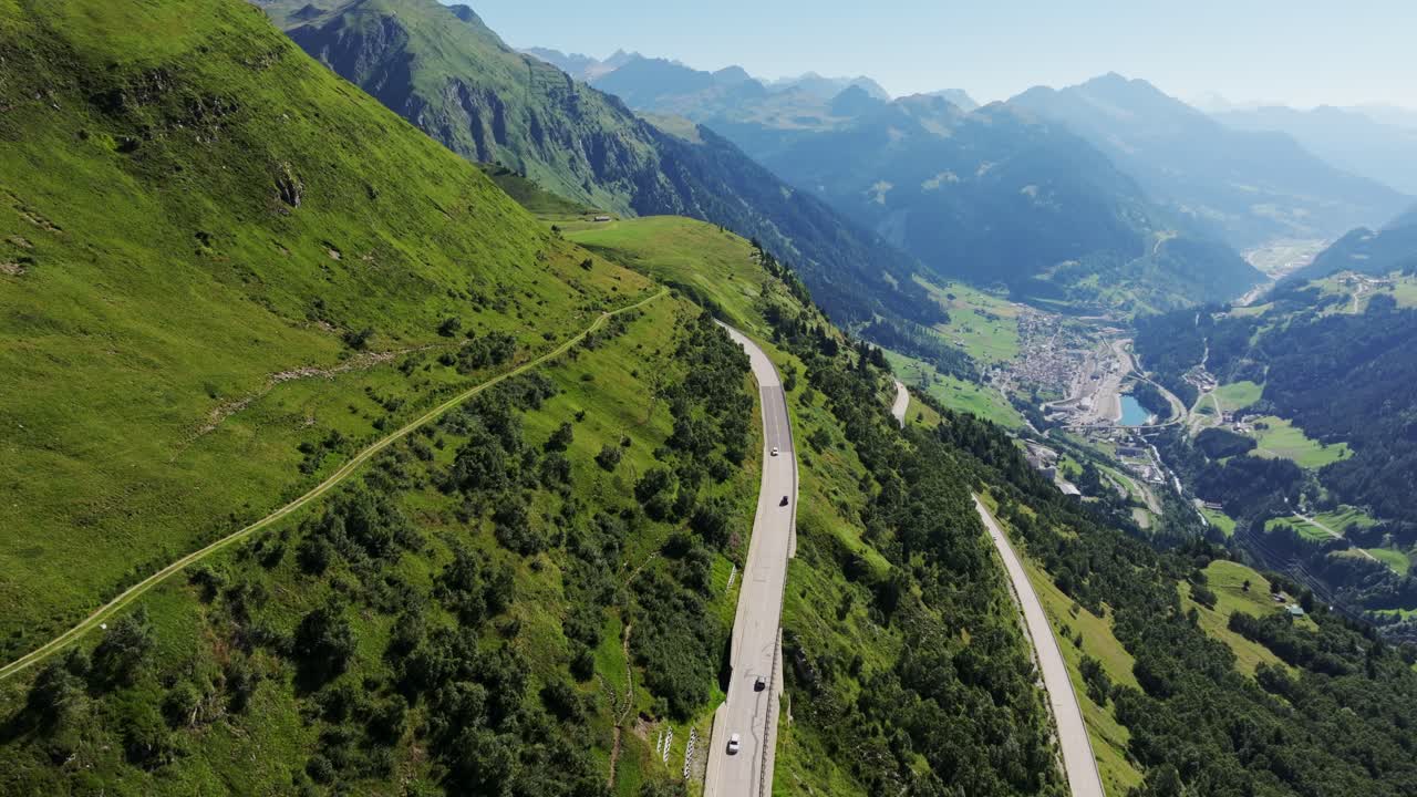 Gotthard Pass road flows through green terrain with high peaks and distant town