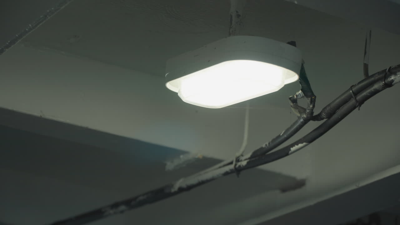 Bright LED ceiling light in garage, exposed wiring and conduit, coolwhite glow casting soft shadows across concrete beams, gritty industrial mood suggesting latenight maintenance or inspection, ideal
