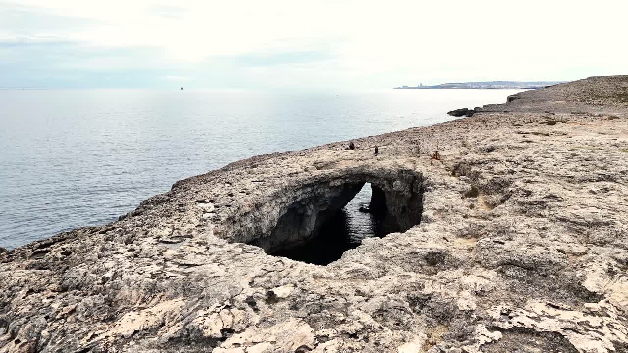 Arco di roccia naturale sul mare a Coral Lagoon a Mellieħa, Malta, con una costa calcarea frastagliata e acqua calma sotto una luce diurna soffusa.