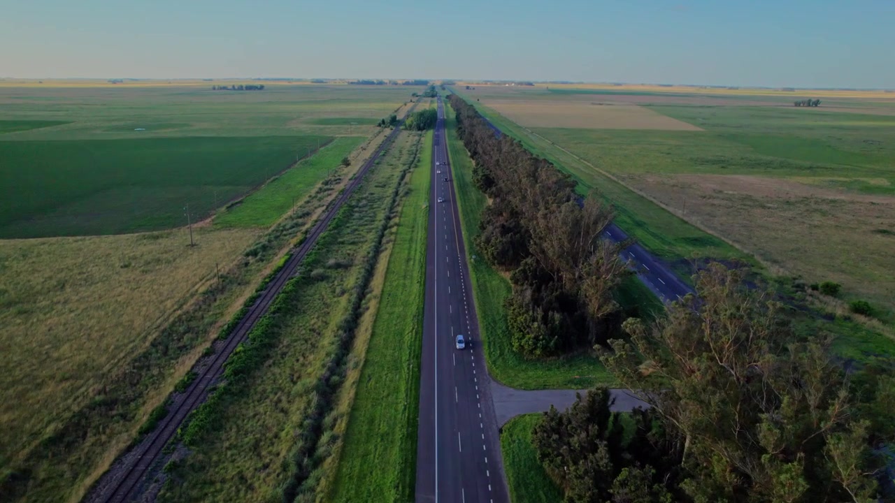 Ujęcie z drona z wysokiej perspektywy, śledzące prostą autostradę z jadącymi samochodami w rozległym wiejskim krajobrazie. Symetryczna kompozycja z zielonymi polami i długimi cieniami.