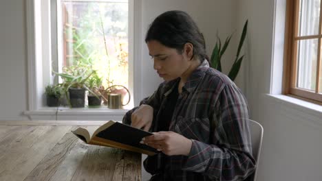 Focused-Puerto-Rican-female-sitting-reading-captivating-story-in-inspirational-book-by-window-light