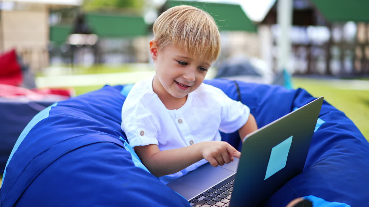 Caucasian six-year old with laptop outdoors. Peaceful boy in bean bag chair outdoors playing computer. Blurred backdrop.
