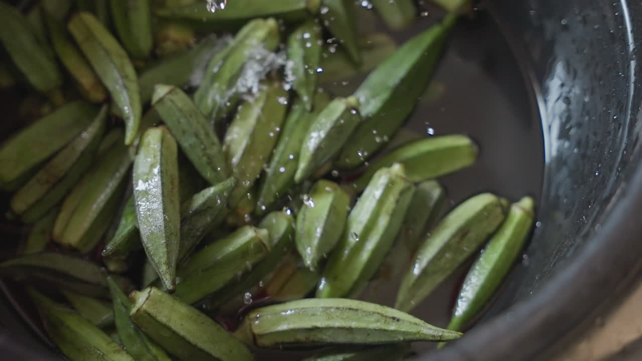 Polong okra segar dibilas dalam mangkuk, tampilan close-up polong hijau mengambang di air dangkal dengan tuangan lembut, tetesan halus di kulit, mangkuk hitam pedesaan di bawah cahaya dapur alami, urutan persiapan makanan.