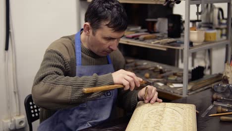 a craftsman in blue overalls carving wooden ornament on backgammon sitting in his working place. portrait of a man working on details using small wooden stick and hammer. own working place