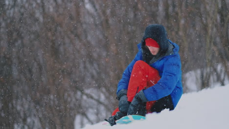 a man in red pants sitting on the snow fastens snowboard shoes and a blue jacket on the ski slope