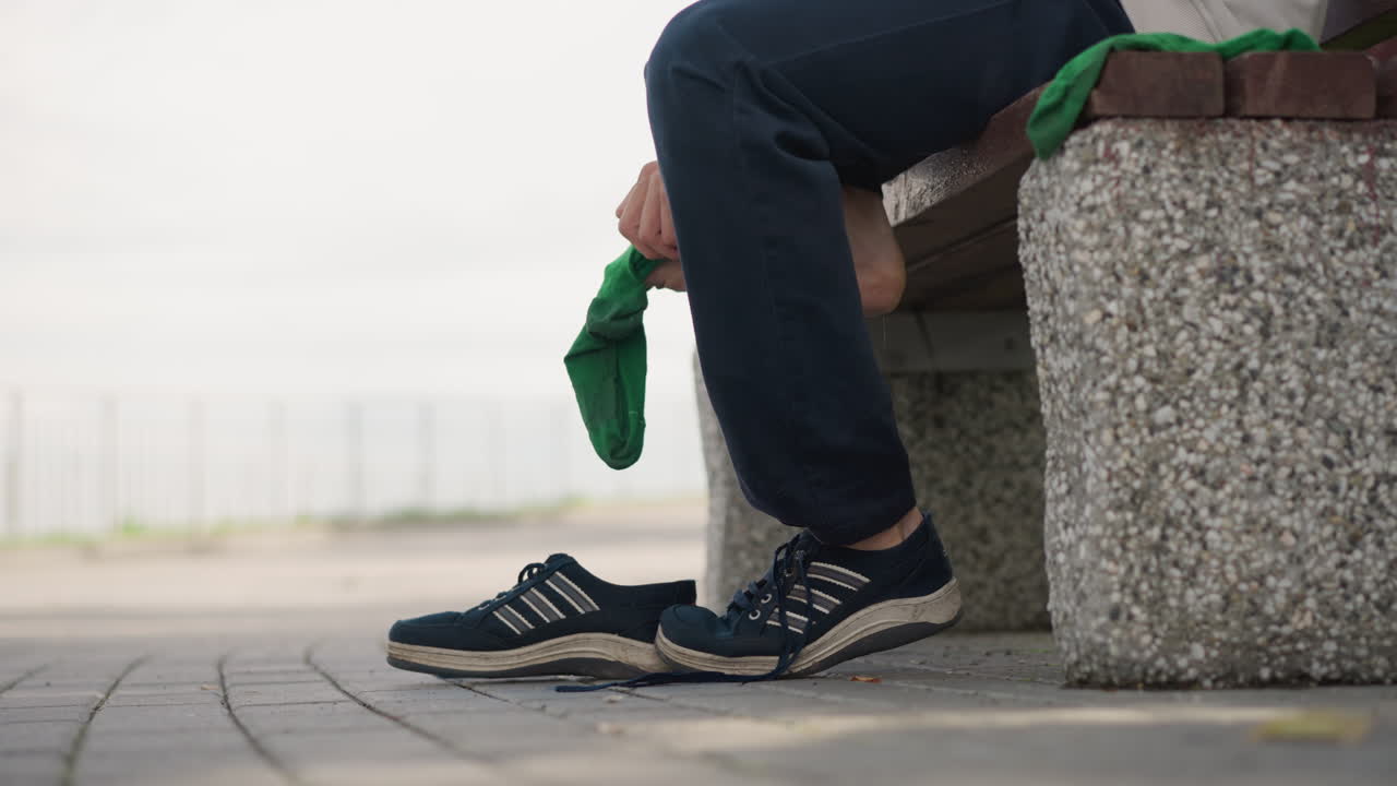 젖은 바닥에서 신발 끈을 묶는 로우 앵글 샷, 콘크리트 벤치에 앉아 운동화 끈을 집중해서 묶는 사람, 근처에 드리워진 초록색 스카프, 워터프런트 울타리와 흐린 하늘이 배경, 클로즈업