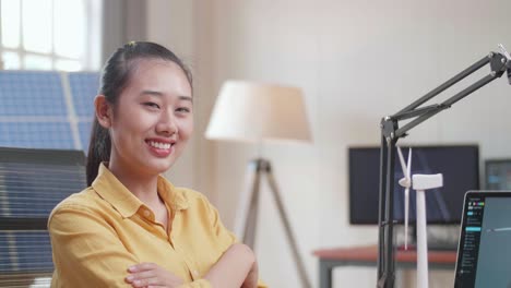 아시아 여성이 풍력 터빈을 바라보고, 팔을 고, 사무실에서 태양전지 에 노트북을 가지고 일하면서 카메라에 미소를 짓고 있습니다.