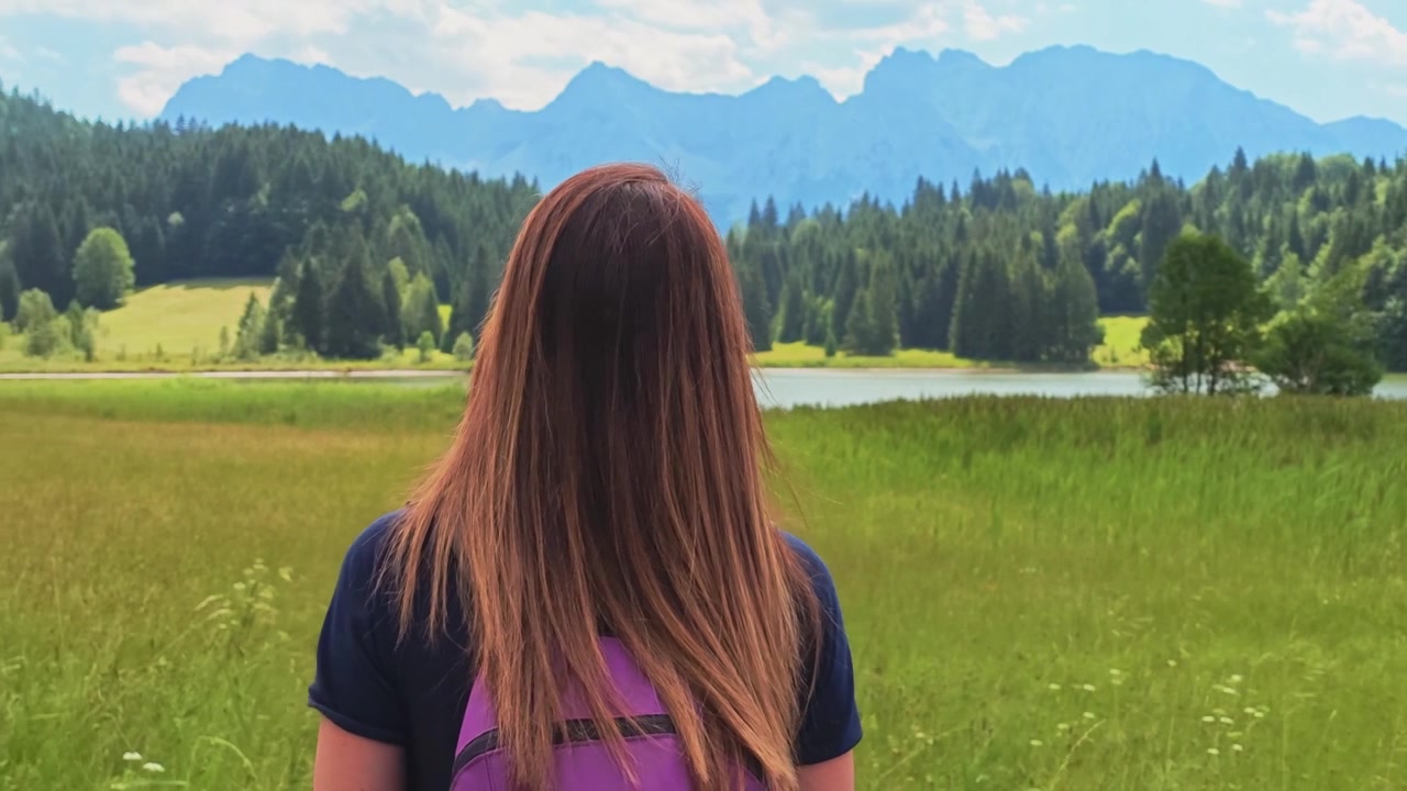 Woman from behind standing in a green meadow. Admiring the scenic view of the Geroldsee and the Bavarian Alps on a sunny day