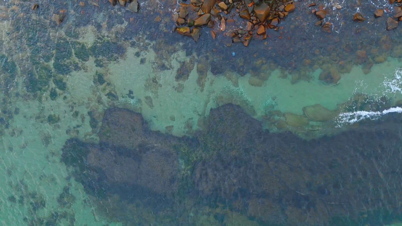 Aerial top view of rocky shoreline and turquoise sea water. Direct overhead drone perspective of dark coastal rocks meeting clear turquoise ocean water with white foam
