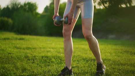 una mujer joven en ropa deportiva se inclina hacia adelante con pesas en la naturaleza en un parque al atardecer. entrenamiento. trabajar en un cuerpo hermoso por la mañana o al atarrear el sol