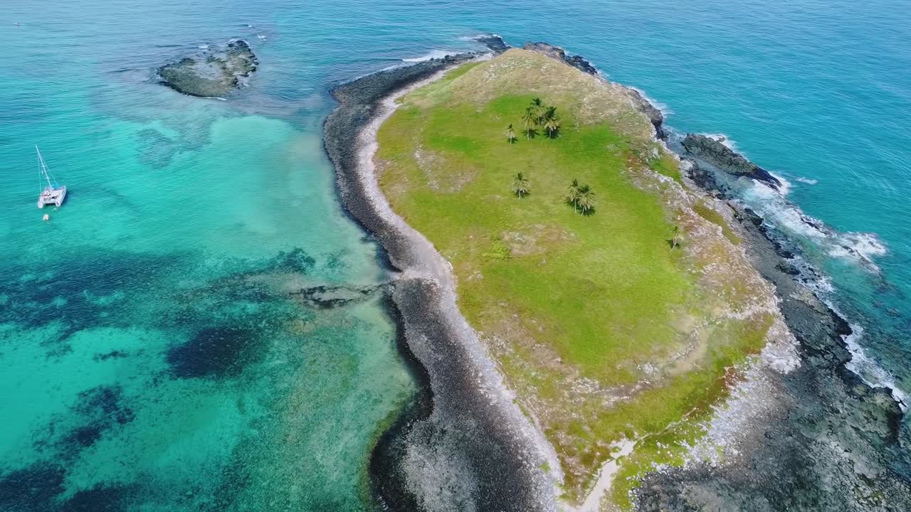 Insel des Abrolhos-Archipels in Bahia, Brasilien, mit leuchtend türkisfarbenem Meerwasser, das eine üppig grüne Landmasse umgibt, und einem segelnden weißen Katamaran, Vorwärtsüberflug und Gierbewegung.