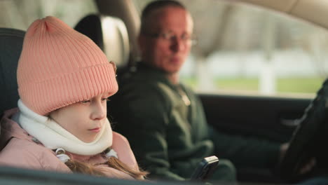 close-up of a young girl in a pink cap and jacket, sitting quietly in a car. she is focused on a smartphone in her hands, a blurred man, likely her father, sits in the driver's seat, observing her