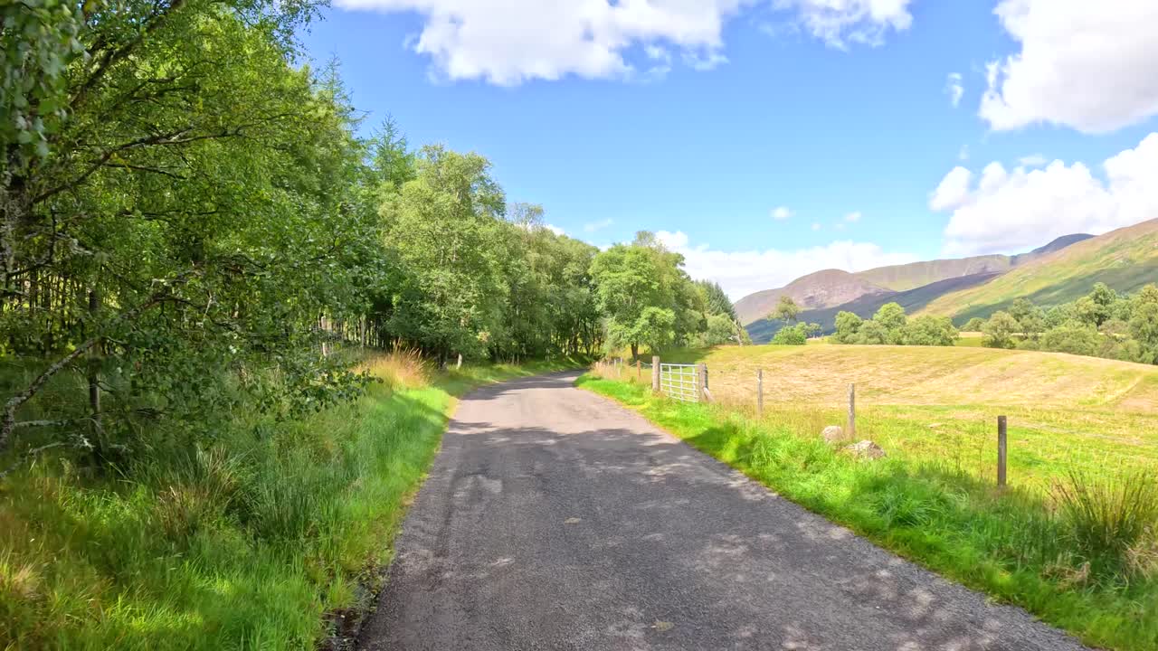 A vehicle travels a narrow country road bordered by trees and grassy fields under bright daylight, with rolling hills and blue sky in the background