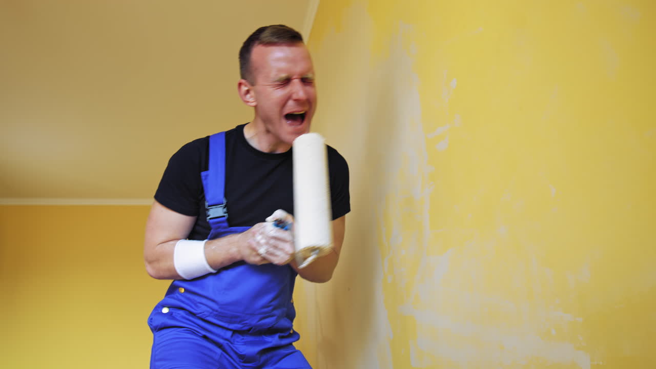 Joyful painter with a paint roller. Funny young man worker in blue overalls singing into a roller while painting walls. Happy moments during makeover in the house.