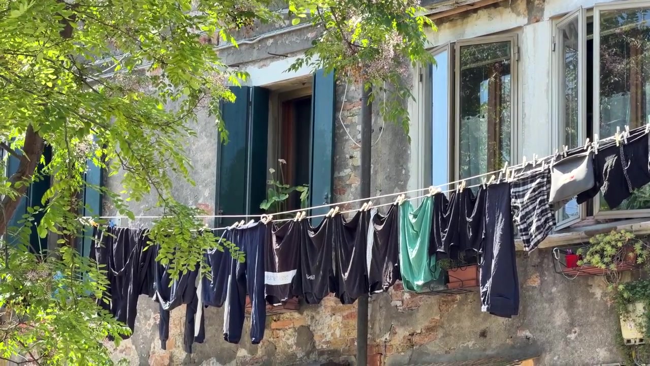 Static 4K shot of laundry drying on a clothesline outside a weathered house. Clothes sway gently in the breeze beneath leafy trees in bright daylight.