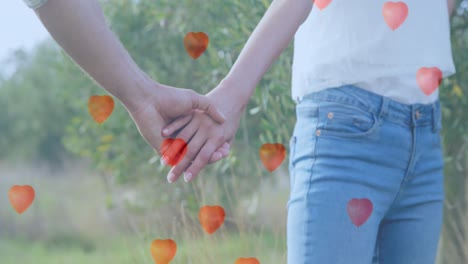 Multiple-heart-balloons-floating-against-mid-section-of-couple-holding-hands-in-the-garden