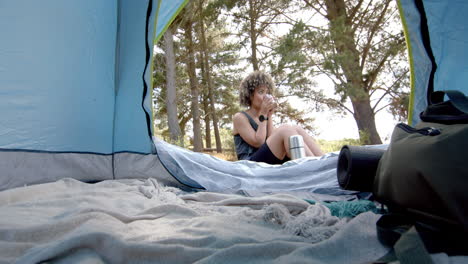 a young biracial woman sits inside a tent, sipping from a mug