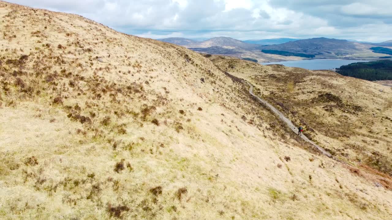 Aerial drone footage gliding close to grassy yellow and brown Scotland hills during a sunny day while a hiker is visible walking on a lonely road. Lake and mountains visible in the background.