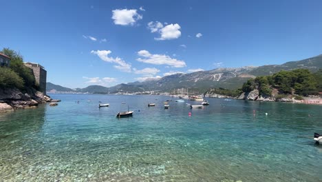 Agua-Azul-Y-Barcos-Que-Rodean-Sveti-Stefan-En-Montenegro-Con-Altas-Montañas-En-El-Fondo,-Playa-Muy-Famosa-Donde-Van-Todas-Las-Celebridades-Actuales-Cuando-Quieren-Pasar-Unas-Vacaciones-Encantadoras