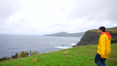 Hombre-Con-Impermeable-Amarillo-Explorando-Acantilados-Volcánicos-Cubiertos-De-Hierba-Junto-Al-Atlántico-En-Castelo-Branco,-Faial