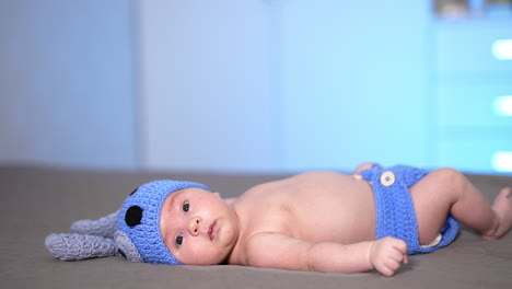 toddler in a cute knitted cap with ears and diaper lying on the bed. sweet child looking attentively at the camera. side view.