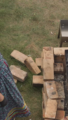 rustic woman in headscarf and traditional clothes watches over fire in brick-lined brazier. female peasant cooks food outdoors on warm summer day upper view