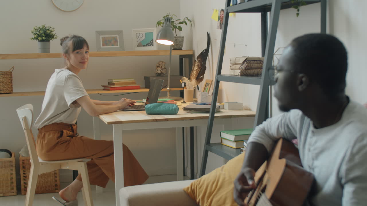 mujer trabajando en una computadora portátil mientras su marido toca la guitarra en casa